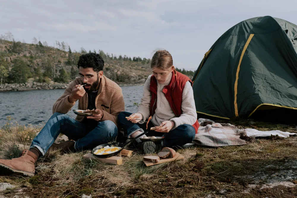 Side view of cheerful young multiracial couple in casual clothes putting scrambled eggs from skillet on plate standing near metal stove in camp