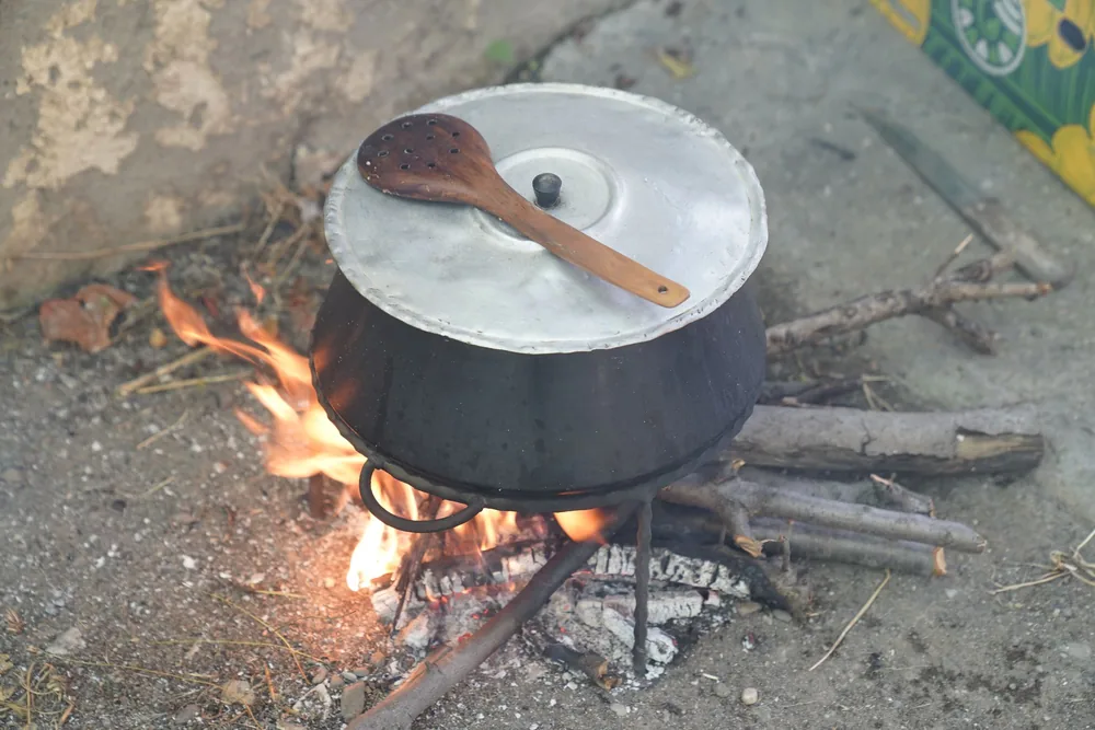 A metal pot with lid sits on an open fire outdoors, with a wooden spoon on top