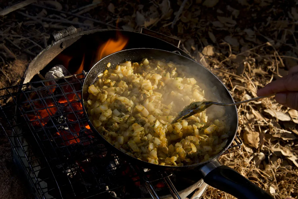 Rustic hash browns cooking on a campfire grill outdoors, emitting steam and aroma