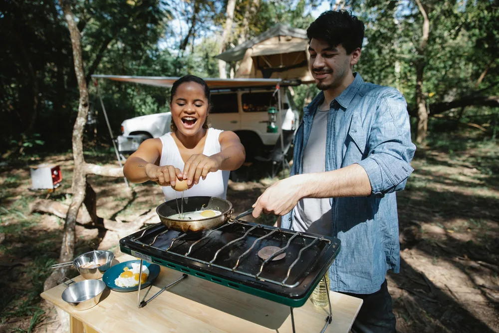 High angle of positive multiracial couple frying eggs on skillet using metal gas stove on wooden table near car and tent in forest