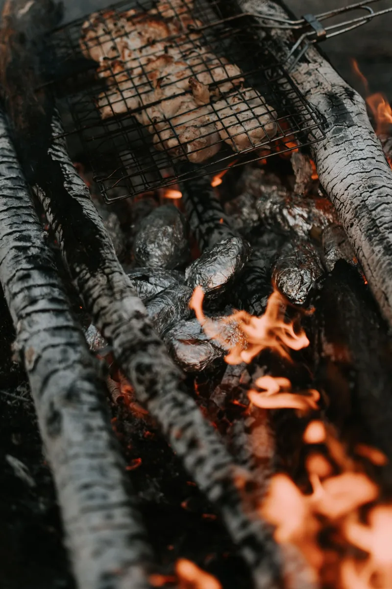 A warm close-up of grilling meat over a campfire with foil-wrapped vegetables, perfect for outdoor cooking enthusiasts