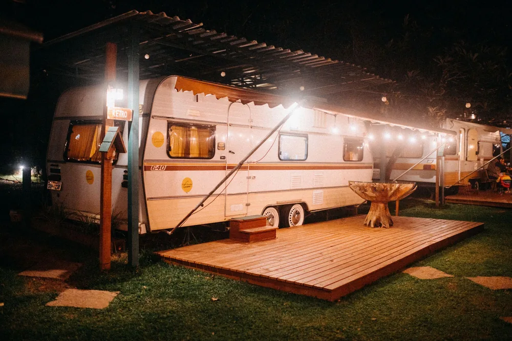 White camper van parked under roof with lights near wooden platform on in trailer park at night