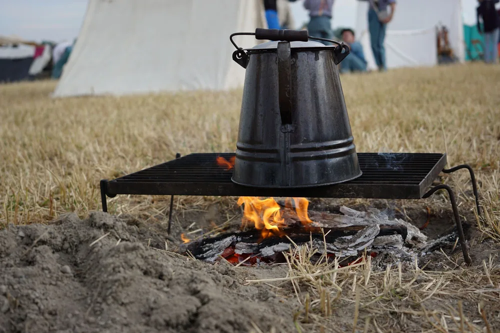 A metal coffee pot heats over an open fire outdoors, evoking a camping adventure