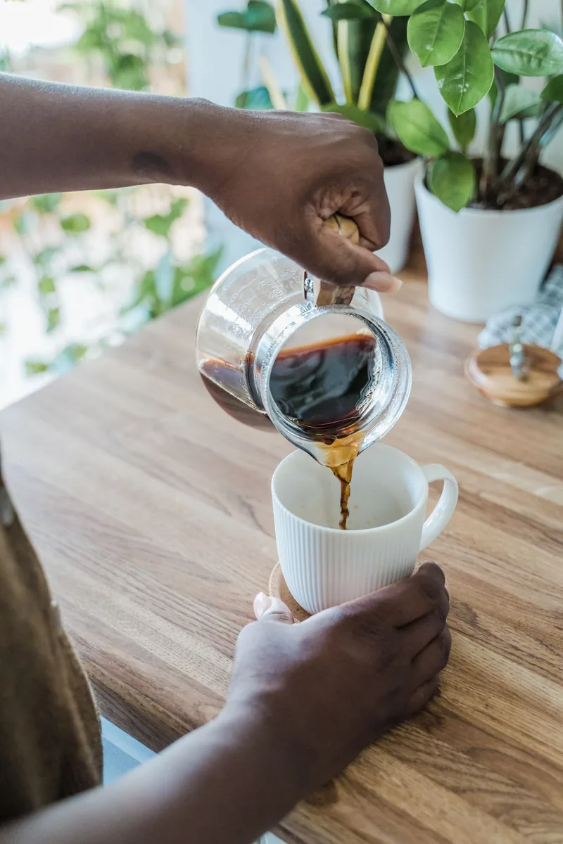 Close-up of hands pouring black coffee from glass pot into mug on a wooden kitchen counter