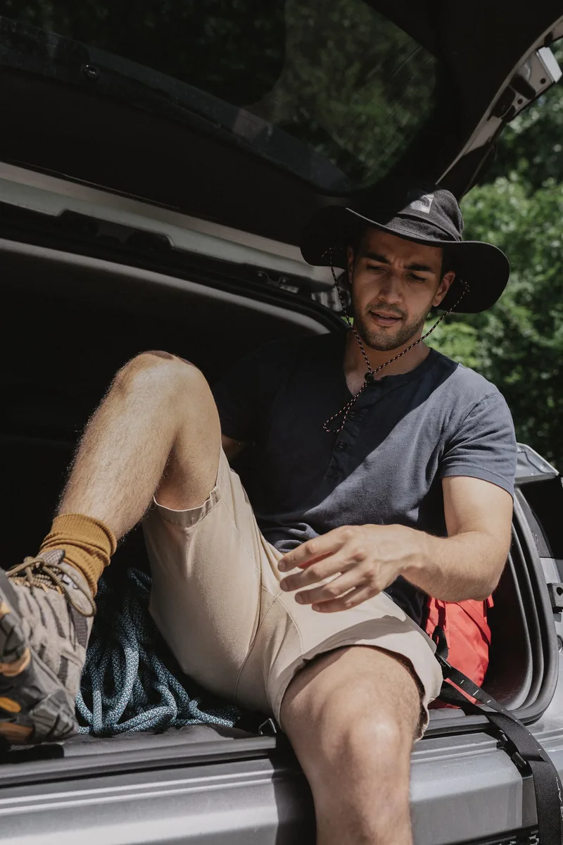 Man sitting in a car trunk, wearing a hat, outdoors with travel gear, enjoying leisure time